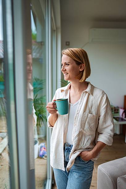 istock Smiling Woman Drinking Coffee and Looking Through Window at Home 2220079451