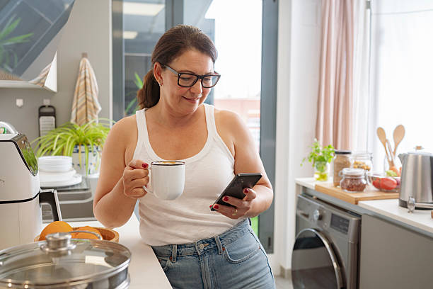 istock Coffee break in the kitchen. Mature woman using phone, scrolling or text messaging. Mobile App. Recipe or online cooking class, Health and wellness 2220080388