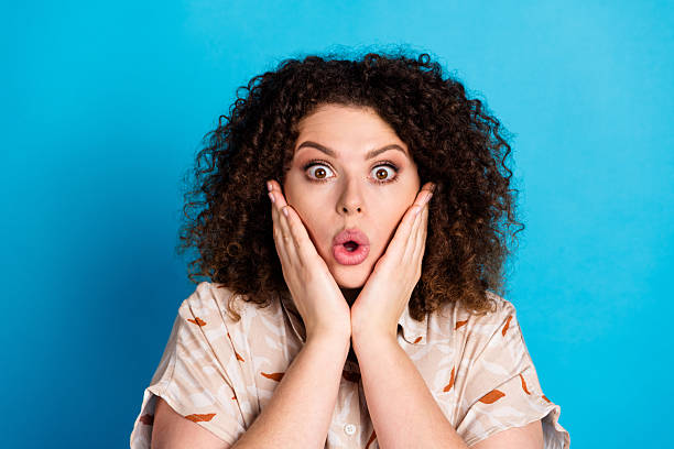 istock Surprised young woman with a vibrant expression displays amazement while touching her cheeks on a bright blue background. 2220082493
