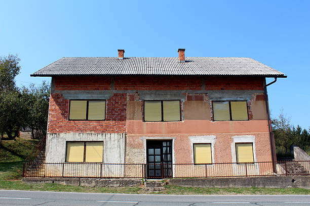Two-story unfinished brick house with yellow shutters and concrete details stands beside a road, showing exposed masonry and a sloped yard with trees under a clear blue sky.