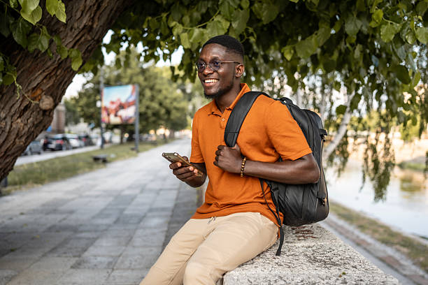 istock Smiling Young Man Outdoors Using Smartphone and Wearing Orange Shirt with Backpack 2220116504