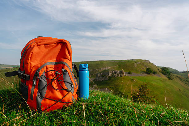 An orange backpack sits on lush green grass next to a blue water bottle, with rolling hills and a cloudy sky in the background during a bright afternoon.