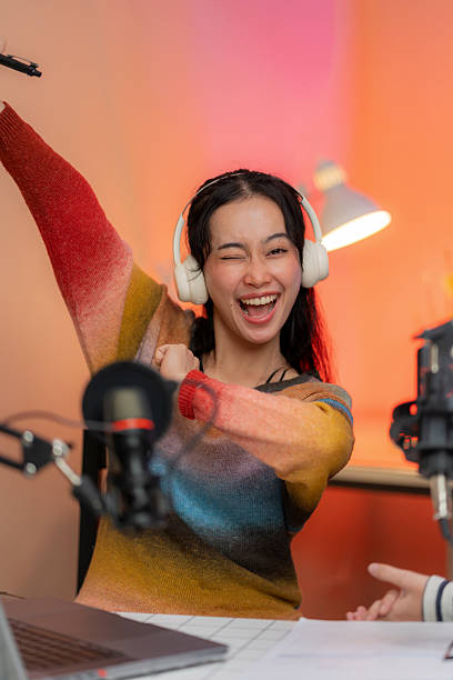 A cheerful podcast presenter celebrates gaining more followers with a bright smile and raised hand while speaking into a microphone. The scene captures the joy of digital success and engaging with audiences in a modern studio setup.