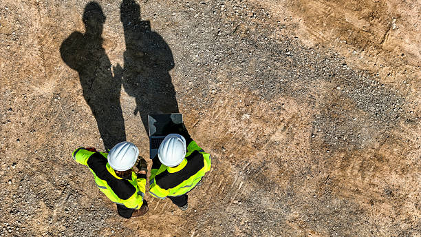 istock Top-down view of two engineers wearing safety vests and helmets using a laptop on a construction site, with their long shadows cast on the ground. 2220121516