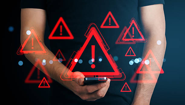 istock Red warning exclamation icons floating around a smartphone held by a person, symbolizing digital alerts and cyber danger on dark background. 2220122646