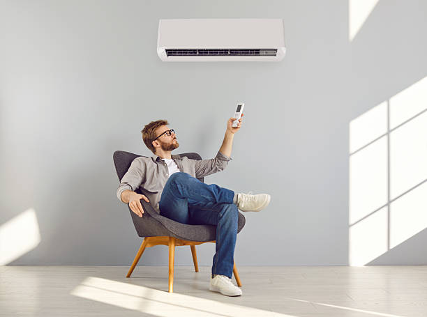 istock Young bearded man turning on air conditioner sitting on chair in a empty space 2220123072