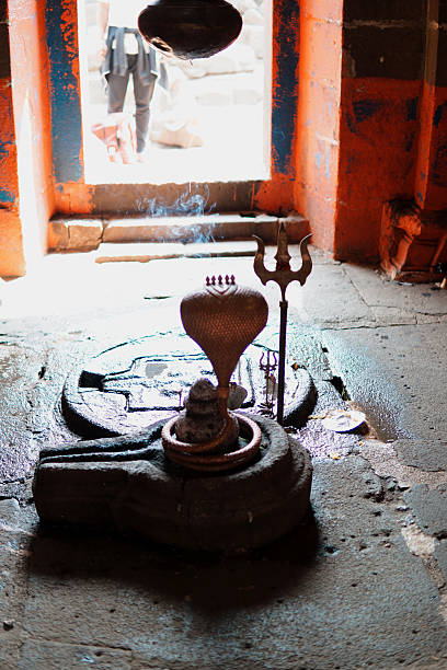 An interior view of a traditional Hindu shrine featuring a sacred Shiva Lingam adorned with a copper serpent hood (Naga) and a trident (Trishul), symbolizing Lord Shiva. Incense smoke gently rises in the dimly lit sanctum, enhancing the serene spiritual ambiance. The temple's aged stone floor and vibrant orange walls frame the scene, offering a glimpse into centuries-old worship practices in India.