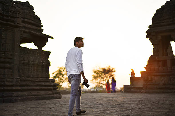 A young man holding a camera strolls through the historic ruins of an ancient temple during golden hour in Maharashtra, India. The silhouettes of weathered stone architecture and the warm hues of the setting sun create a perfect reflective scene.