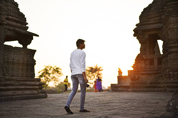 A young man holding a camera strolls through the historic ruins of an ancient temple during golden hour in Maharashtra, India. The silhouettes of weathered stone architecture and the warm hues of the setting sun create a perfect reflective scene.