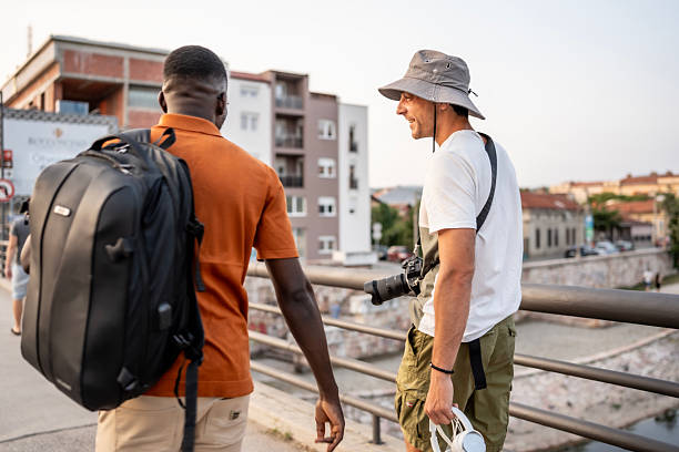 Two individuals walk across a bridge in an urban setting, engaging in friendly discussion while carrying a camera and a backpack, conveying exploration, companionship, and outdoor adventure.