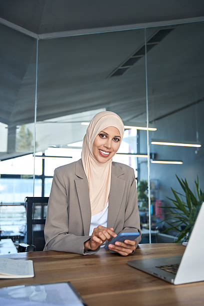 istock Middle Eastern muslim businesswoman manager ceo using cell phone mobile app. Smiling young indian woman in hijab holding smartphone sitting in office working. Vertical portrait looking at camera 2220138155