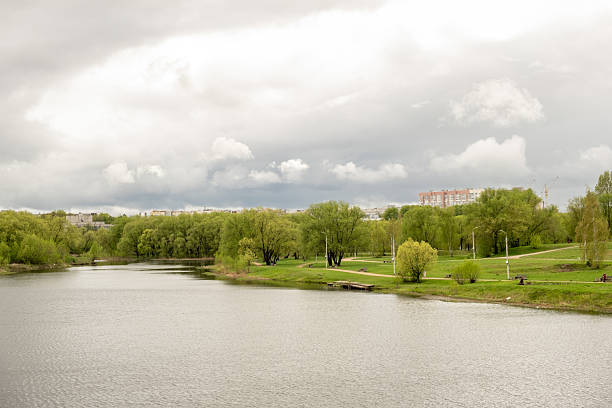 A serene river flows through a vibrant park filled with lush trees, surrounded by gently rolling land under an overcast sky in the spring season.