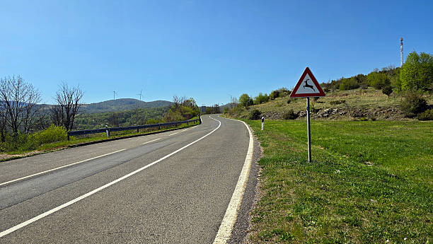 Curving asphalt road bordered by grassy slopes and forest, marked with a triangular deer crossing warning sign; wind turbines and a cell tower visible on the green hills under a clear blue sky