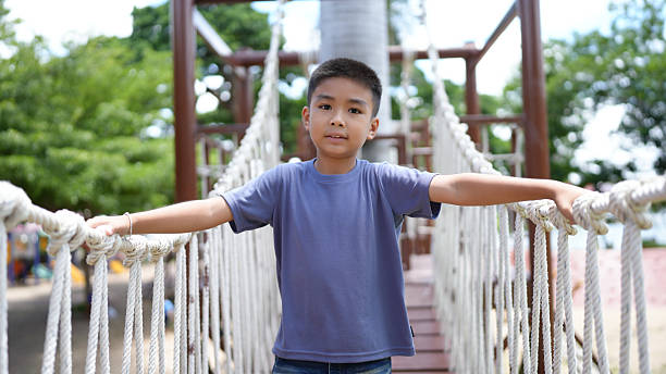 istock Young boy stands confidently on rope bridge in playground, surrounded by greenery and bright skies. His expression reflects joy and curiosity as he enjoys his time outdoors 2220157584