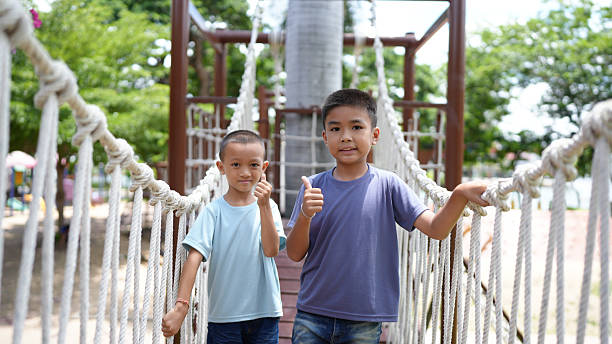 istock Two boys smiling and giving thumbs up while standing on rope bridge in playground, surrounded by greenery and colorful play equipment, showcasing joy and friendship 2220157587
