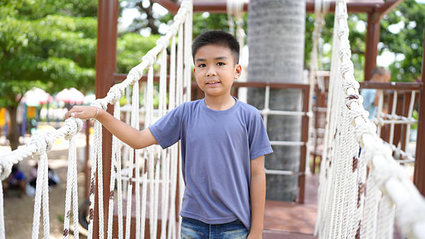Young boy stands confidently on rope bridge in park, surrounded by greenery and playful atmosphere. His casual outfit and cheerful demeanor reflect sense of adventure and joy