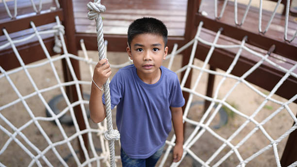 Young boy stands confidently playground structure, gripping rope. His expression is curious and focused, surrounded by net like climbing frame. scene captures moment of childhood adventure