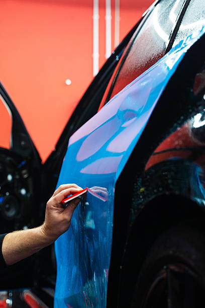 istock Vehicle wrap detailing involves squeegee strokes for seamless, wrinkle-free adhesion. Detailing expert uses red squeegee wrapper to flatten blue PPF wrap on car surface. 2220160060