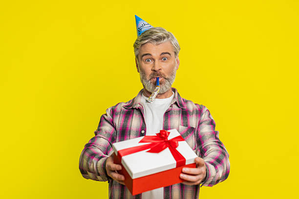 istock Mature man handing presenting birthday gift box cone hat blower joyful expression festive atmosphere 2220169298