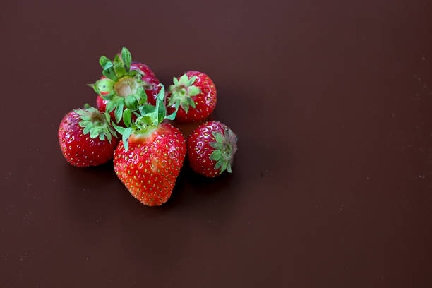Red strawberries with green stems form a cluster on a dark brown background. This composition highlights the freshness and vibrancy of the berries against a neutral, dark backdrop.