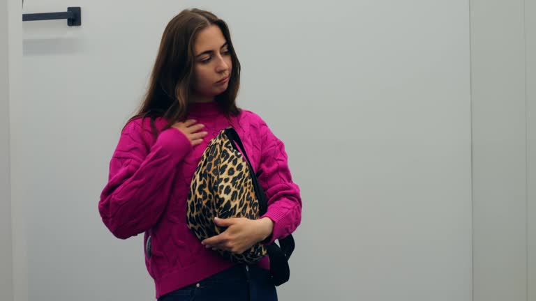 Young woman selecting leopard print accessory, wearing fuchsia cable knit sweater while choosing matching bag inside clothing store dressing room. Concept of buying women clothes