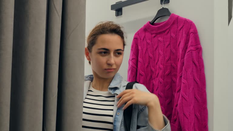 Young muslim woman wearing hijab trying on clothes in a dressing room, matching pink sweater with leopard print bag and looking at herself in the mirror. Woman sheds hijab and puts on stylish clothes