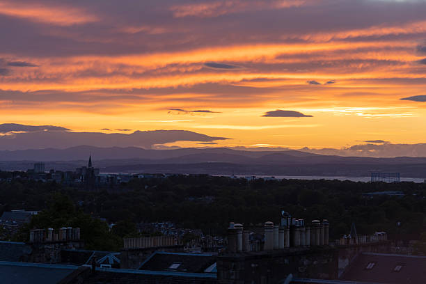 A breathtaking view of Edinburgh's city skyline during a colorful sunrise and the blue hour, captured from Calton Hill. The iconic Balmoral Clock Tower glows warmly amid the vibrant streets and traffic below. Surrounded by colorful autumn trees, this scenic vista highlights the charm of Scotland’s capital city in the fall.