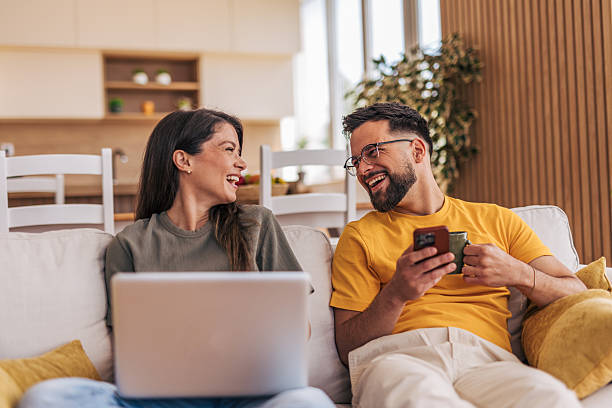 istock Young couple relaxing on sofa, using laptop and smartphone, laughing together 2220220286