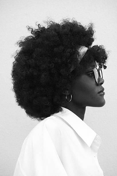 A striking black-and-white portrait captures a woman in profile, her gaze steady and composed. Her natural curls form a soft halo, contrasted by the structured white shirt and dark glasses. The clean background focuses all attention on her presence - calm, intellectual, quietly powerful. The simplicity amplifies her individuality and self-possession.