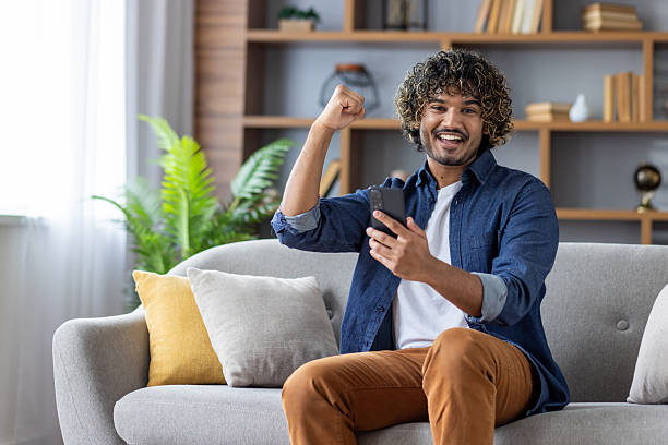 istock Excited man with curly hair celebrates success while using a smartphone at home on the sofa. 2220226264