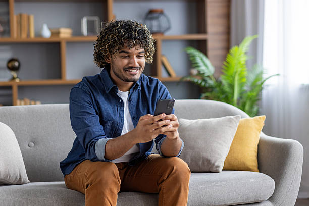 istock Young successful man using app on phone while sitting inside apartment on sofa at home. Satisfied smiling typing text message and browsing social media 2220226265