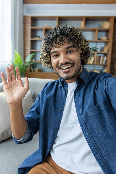 istock Vertical shot, man smiling and looking at camera, waving hello, using app on phone for video call. Sitting at home in living room of apartment 2220226283