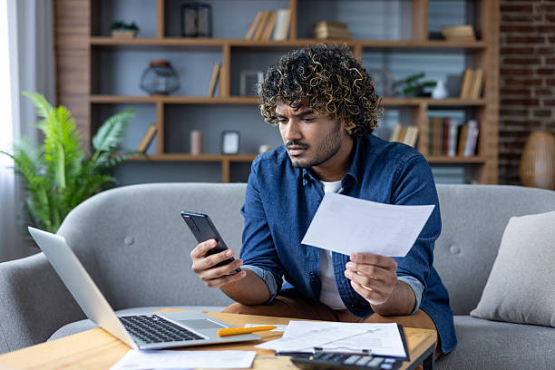 istock Young man looking worried using a smartphone while checking bills at his home office. 2220226338