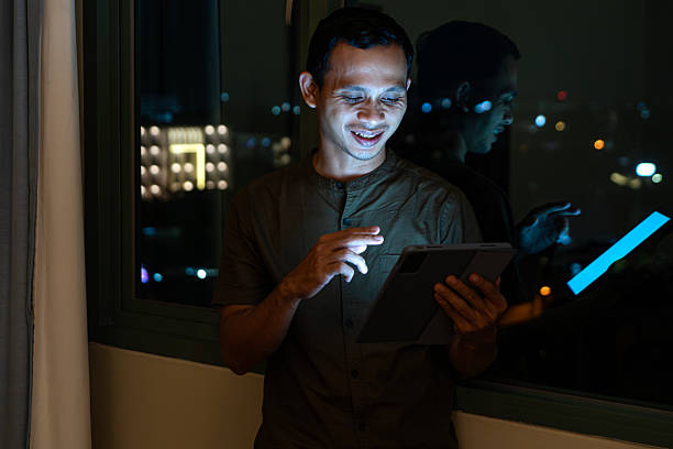Young asian man smiling while using a tablet near the window at night, screen glow illuminating his face with city lights reflection.