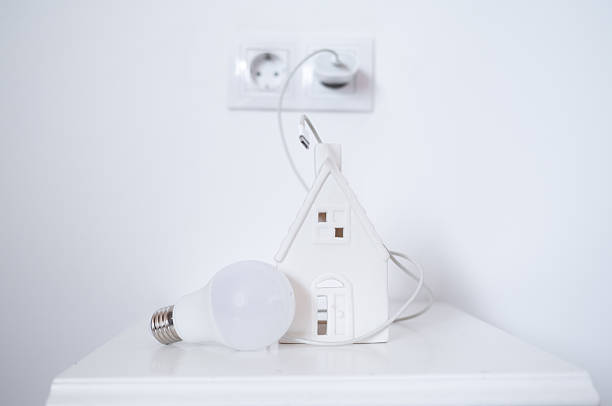 Miniature house is connected to a power outlet with a cable, symbolizing energy consumption, while an led light bulb lies nearby, representing energy-efficient lighting solutions for homes