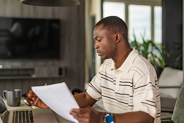 istock Young african businessman sitting at a table at home working on a laptop and writing notes down on a notepad 2220233022