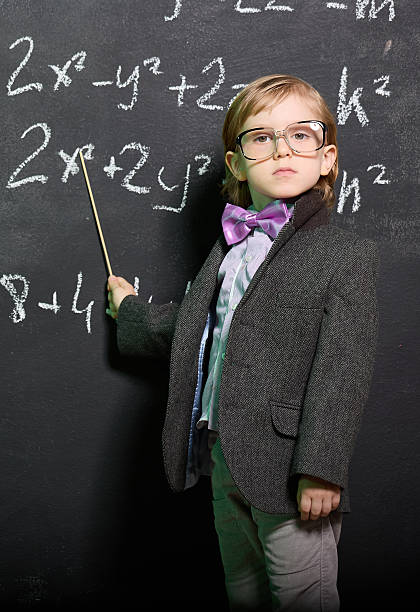 A cute little boy with big glasses, dressed in a suit and a bow tie, stands at a chalkboard covered with mathematical symbols and formulas. The boy is pointing at the formulas on the board, looking over his shoulder at the camera