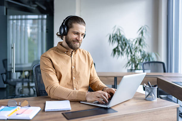 istock Bearded man wearing headphones working on a laptop in modern office environment, looking happy. 2220233721