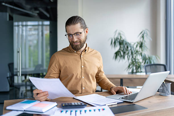 istock Confident Professional Reviewing Documents and Working on a Laptop in Modern Office 2220233748