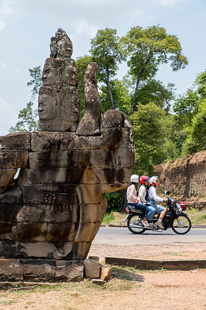 Siem Reap, Cambodia - April 6, 2025: Sculptures at the South Gate of Angkor Thom and a motorcycle with three passengers in Siem Reap, Cambodia