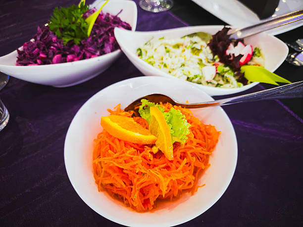 Close-up of various fresh vegetable salads served in white bowls, including grated carrot with orange, red cabbage, and cucumber salad with radish, on a banquet table with purple table runner.