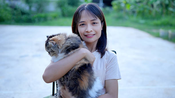 A young woman smiles while holding fluffy cat in her arms, showcasing warm bond between them. background features greenery, adding serene atmosphere