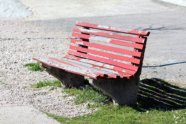 Old red park bench with peeling paint and concrete supports sits on gravel and grass in an outdoor public area, showing signs of age and use in bright sunlight