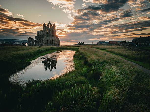 A dramatic view of the iconic Whitby Abbey ruins silhouetted against a vivid sunset sky in North Yorkshire, England. This ancient Gothic structure stands atop the East Cliff, casting its reflection in a stagnant pool on the moor, surrounded by wild grasses. The warm golden light of dusk highlights the texture of the stonework while clouds streak the sky with rich hues of orange, purple, and blue. This atmospheric scene captures the haunting beauty and significance of this world famous medieval monastery, which inspired Bram Stoker's Dracula and overlooks the picturesque coastal town of Whitby.