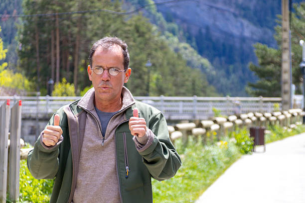 A middle-aged man, dressed in a green jacket, stands outdoors giving thumbs up with a mountainous backdrop. The setting is bright and sunny, capturing a joyful mood in nature.