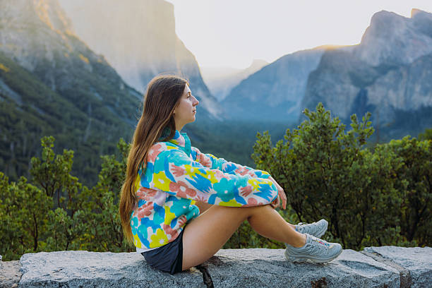 Side view of female with long hair in colourful fleece shirt sitting at the scenic viewpoint of summer sunrise above majestic mountain range with forest and waterfall in Yosemite National park, California, the United States