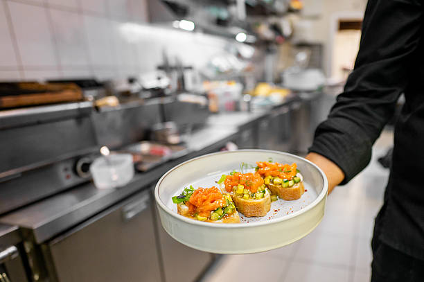 A chef holds a plate with three bruschetta topped with diced vegetables and herbs in a professional kitchen. The background shows various kitchen equipment, emphasizing a busy culinary setting
