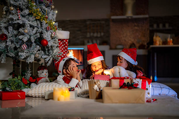 istock Three kids in Santa hats gather under a Christmas tree, surrounded by glowing presents and cozy decorations. They open gift boxes and share festive joy in a warm holiday setting. 2220264634