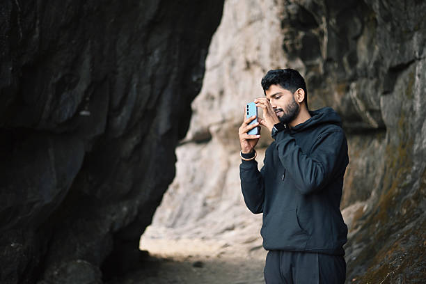 Inside a narrow rocky canyon, at Ghangad fort, Akola a young man dressed in a black hoodie stands focused, holding up his phone to frame the perfect picture. Surrounded by towering rock formations and textured stone walls, his expression shows concentration and appreciation for the rugged beauty around him.