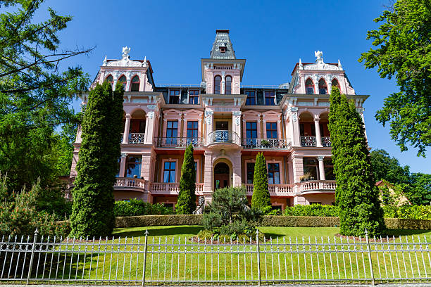 Heringsdorf, Mecklenburg-Western Pomerania, Germany - May 29, 2025: Beautifully restored historic villa in Heringsdorf with pink exterior, balconies, and manicured garden under clear blue sky.